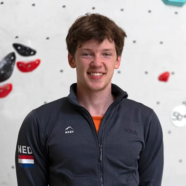 Een jonge man met kort bruin haar glimlacht naar de camera; hij draagt een donker jack met ritssluiting en een patch met de Nederlandse vlag. Hij staat voor een klimwand binnenshuis met rode en zwarte